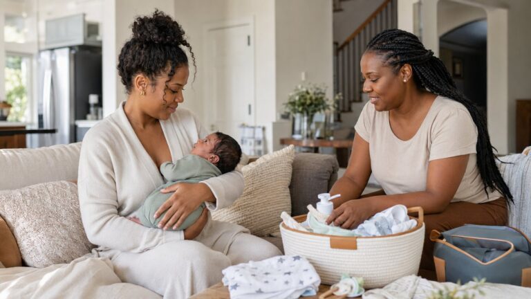 A mother holding her newborn while a postpartum caregiver organizes baby supplies nearby